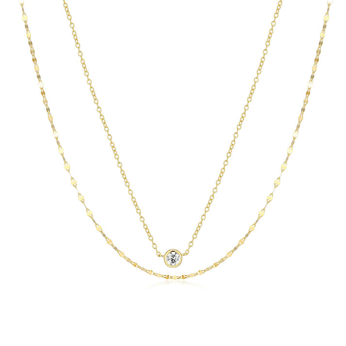 A double strand gold necklace with a bezel set clear stone on the shorter of the two chains on a white background